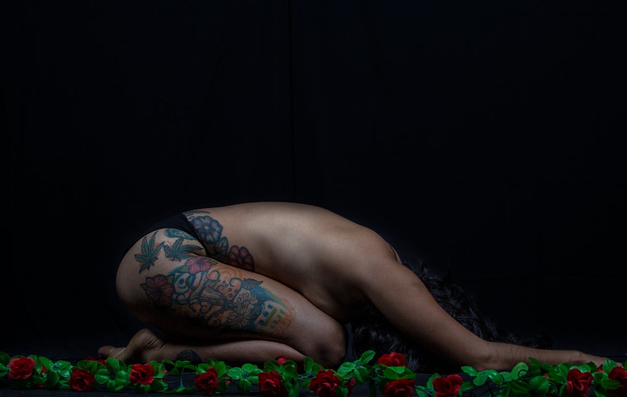 Artistic nude woman with tattoos in yoga pose surrounded by roses against black background.